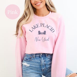 May include: A light pink sweatshirt with the text "Lake Placid New York" in navy blue. The design includes two small chair illustrations. The sweatshirt is a long-sleeved crew neck.