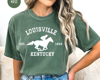 May include: Moss green t-shirt with a white graphic of a horse and jockey, and the text "LOUISVILLE KENTUCKY EST. 1828". The shirt is a relaxed fit and has a vintage look.