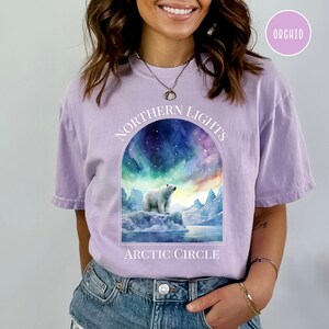 Op de afbeelding: Lichtpaarse t-shirt met een afbeelding van een ijsbeer op een ijsberg onder het noorderlicht. De tekst "NORTHERN LIGHTS" staat boven de afbeelding en "ARCTIC CIRCLE" eronder. Het shirt is effen van kleur.
