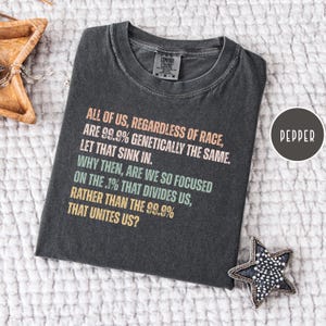 May include: Dark grey t-shirt with the text "ALL OF US, REGARDLESS OF RACE, ARE 99.9% GENETICALLY THE SAME. LET THAT SINK IN." The shirt also has the text "WHY THEN, ARE WE SO FOCUSED ON THE .1% THAT DIVIDES US, RATHER THAN THE 99.9% THAT UNITES US?"