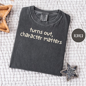 May include: A charcoal gray short-sleeve t-shirt with a crew neckline. The shirt features the text "turns out, character matters" printed in a light cream color across the chest. The garment has a visible brand label inside the collar.