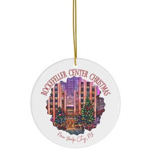 May include: A white ceramic Christmas ornament featuring a colorful image of the Rockefeller Center Christmas tree in New York City. The ornament has a red glitter design with the text "Rockefeller Center Christmas" and "New York City, NY".