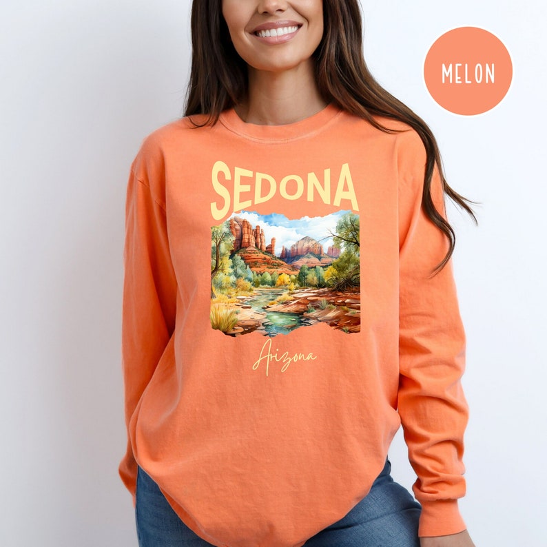 May include: A long-sleeved, orange t-shirt with a graphic print of a landscape scene with red rock formations and a river. The text "SEDONA Arizona" is printed on the shirt.