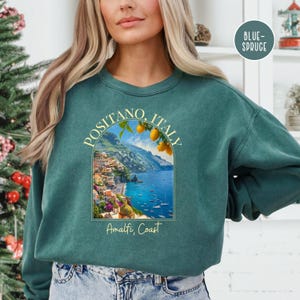 K&ouml;nnte beinhalten: Ein gr&uuml;nes Sweatshirt mit einem Grafikdruck von Positano, Italien, mit einer farbenfrohen Stadtlandschaft, einem blauen Meer und Zitronenb&auml;umen. Der Text "Positano, Italien" und "Amalfik&uuml;ste" ist auf dem Sweatshirt aufgedruckt.