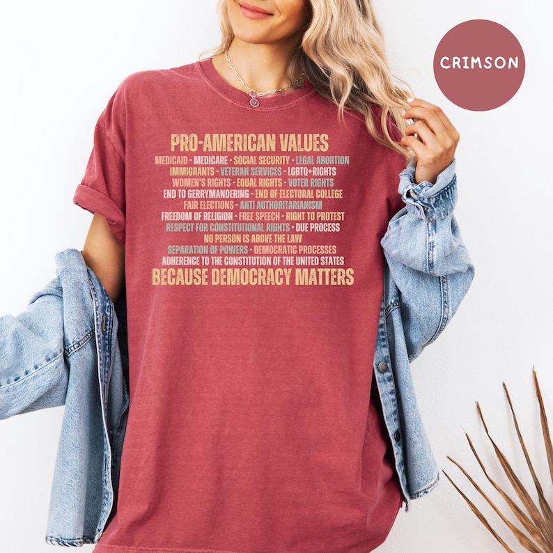 Pode incluir: Camiseta de cor carmesim com o texto "PRO-AMERICAN VALUES" e uma lista de valores relacionados. A frase "BECAUSE DEMOCRACY MATTERS" est&aacute; na parte inferior. A camisa &eacute; de cor s&oacute;lida e tem um estilo casual.