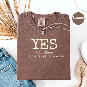 以下が含まれることがあります： 「YES to coffee. no to everything else.」の文字が入った茶色のTシャツ。シルバーのネックレスがTシャツの上に置かれています。Tシャツは、ジーンズと白い靴の隣の白い表面に折りたたまれています。