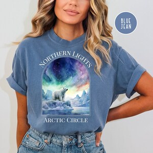 Op de afbeelding: Blauwe spijkerbroekkleurige t-shirt met een aquarel illustratie van een ijsbeer op een ijsberg onder het noorderlicht. De tekst "NORTHERN LIGHTS" staat in een boog boven de afbeelding, met "ARCTIC CIRCLE" eronder. Een kleine cirkel rechtsboven zegt "BLUE JEAN".