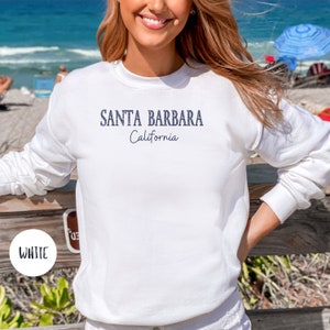 May include: White sweatshirt with the text "SANTA BARBARA California" in navy blue. The sweatshirt is long-sleeved and has a crew neck. The word "WHITE" is in a white circle in the bottom left corner.