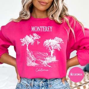 May include: A pink sweatshirt with a white graphic design of trees and the text "Monterey California". The text "Heliconia" is visible in the lower right corner of the image.