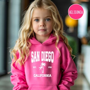 May include: A young person wearing a bright pink San Diego hoodie. The hoodie features the city name in bold white letters, along with a palm tree graphic and the text "California" and "Est. 1769". The hoodie has a hood and a front pocket.