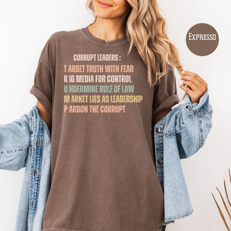 May include: A brown t-shirt with the text "CORRUPT LEADERS: TARGET TRUTH WITH FEAR, RIG MEDIA FOR CONTROL, UNDERMINE RULE OF LAW, MARKET LIES AS LEADERSHIP, PARDON THE CORRUPT" in various colors. The shirt is worn with a denim jacket.