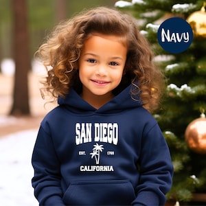 May include: A navy blue hooded sweatshirt featuring the text "SAN DIEGO" in white block letters, with "EST. 1769" and a palm tree graphic below. The word "CALIFORNIA" is printed at the bottom. The sweatshirt is worn by a young person.