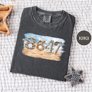May include: Dark gray Comfort Colors t-shirt with a beach-themed graphic. The graphic features the word "86417" spelled out with seashells on a sandy beach with a blue sky background. The shirt is laid flat on a white textured surface.