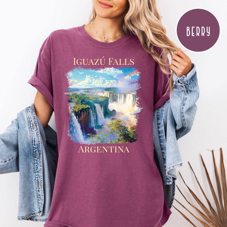 Pu&ograve; includere: T-shirt color bacca con un'immagine panoramica delle cascate dell'Iguaz&uacute; in Argentina. La grafica include il testo "IGUAZ&Uacute; FALLS" sopra le cascate e "ARGENTINA" sotto. La maglietta ha maniche arrotolate e una vestibilit&agrave; rilassata.