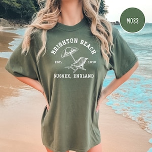 K&ouml;nnte beinhalten: Ein moosgr&uuml;nes T-Shirt mit einem wei&szlig;en Grafikdesign, auf dem "Brighton Beach Est. 1913 Sussex, England" steht. Die Grafik zeigt einen Strandstuhl und einen Sonnenschirm.