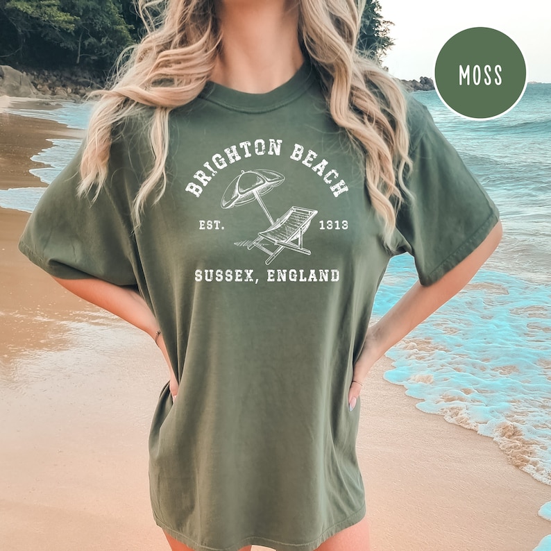 K&ouml;nnte beinhalten: Ein moosgr&uuml;nes T-Shirt mit einem wei&szlig;en Grafikdesign, auf dem "Brighton Beach Est. 1913 Sussex, England" steht. Die Grafik zeigt einen Strandstuhl und einen Sonnenschirm.