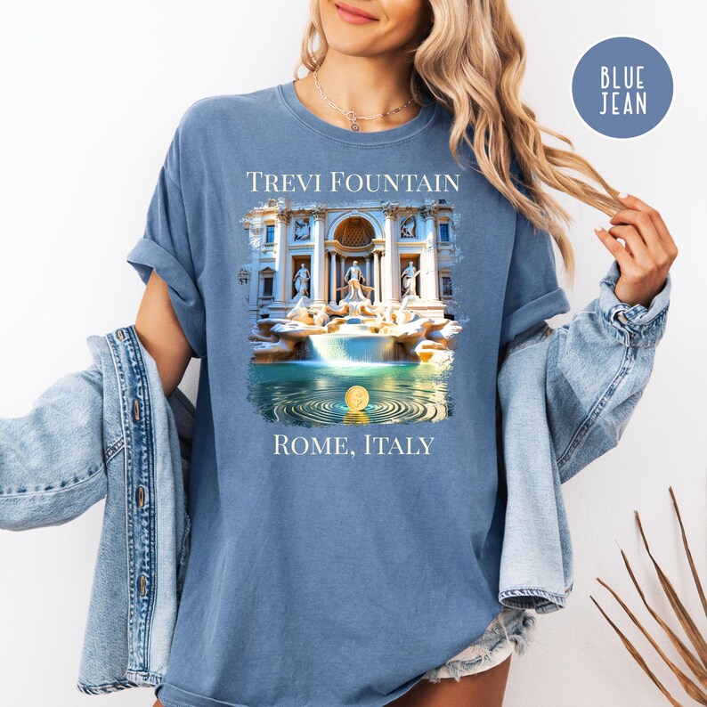May include: A blue t-shirt with a graphic of the Trevi Fountain in Rome, Italy. The text "TREVI FOUNTAIN" is above the image, and "ROME, ITALY" is below. The shirt is a solid blue colour.