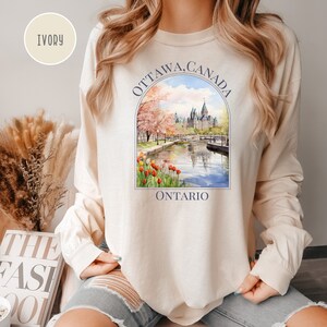 May include: A long-sleeved, ivory-colored sweatshirt with a graphic print of the Parliament Building in Ottawa, Canada. The graphic is surrounded by a blue circle with the text "OTTAWA, CANADA" and "ONTARIO" underneath.