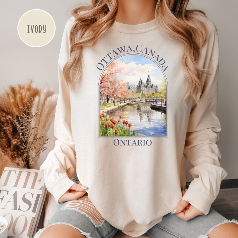 May include: A long-sleeved, ivory-colored sweatshirt with a graphic print of the Parliament Building in Ottawa, Canada. The graphic is surrounded by a blue circle with the text "OTTAWA, CANADA" and "ONTARIO" underneath.