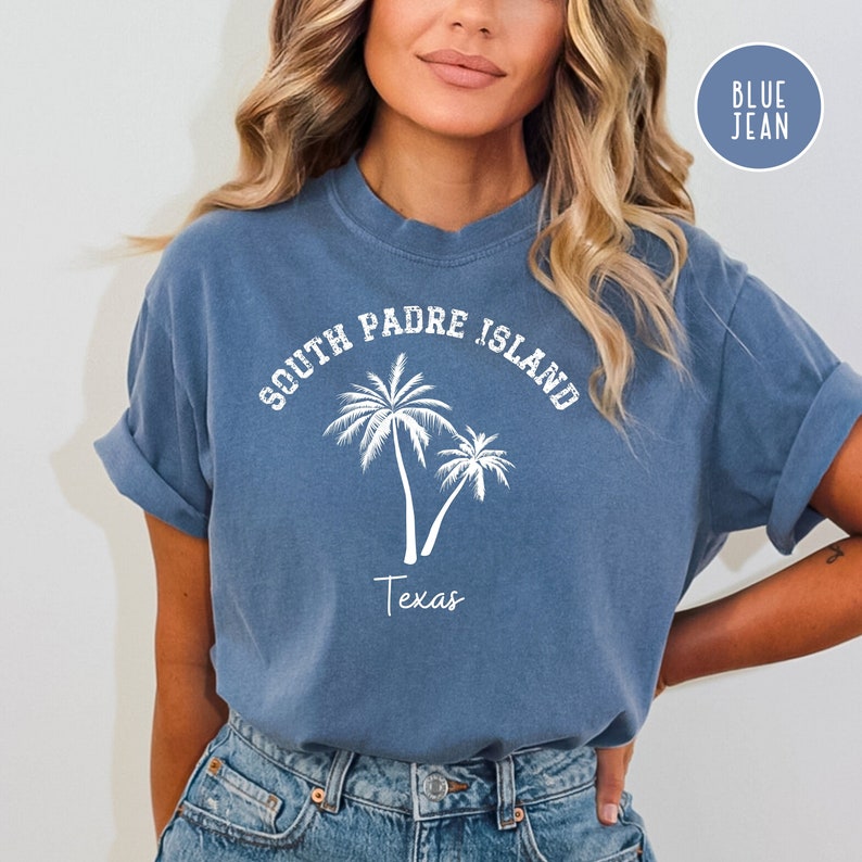 May include: A blue jean t-shirt with white text that reads "South Padre Island Texas" and a graphic of two palm trees.