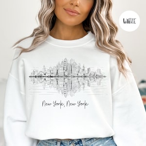 K&ouml;nnte beinhalten: Wei&szlig;er Pullover mit einer schwarz-wei&szlig;en Grafik der Skyline von New York City, die sich im Wasser spiegelt. Der Text "New York, New York" ist unter der Grafik gedruckt.