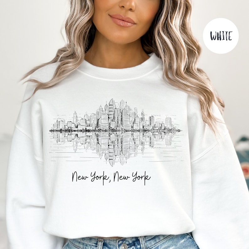 K&ouml;nnte beinhalten: Wei&szlig;er Pullover mit einer schwarz-wei&szlig;en Grafik der Skyline von New York City, die sich im Wasser spiegelt. Der Text "New York, New York" ist unter der Grafik gedruckt.
