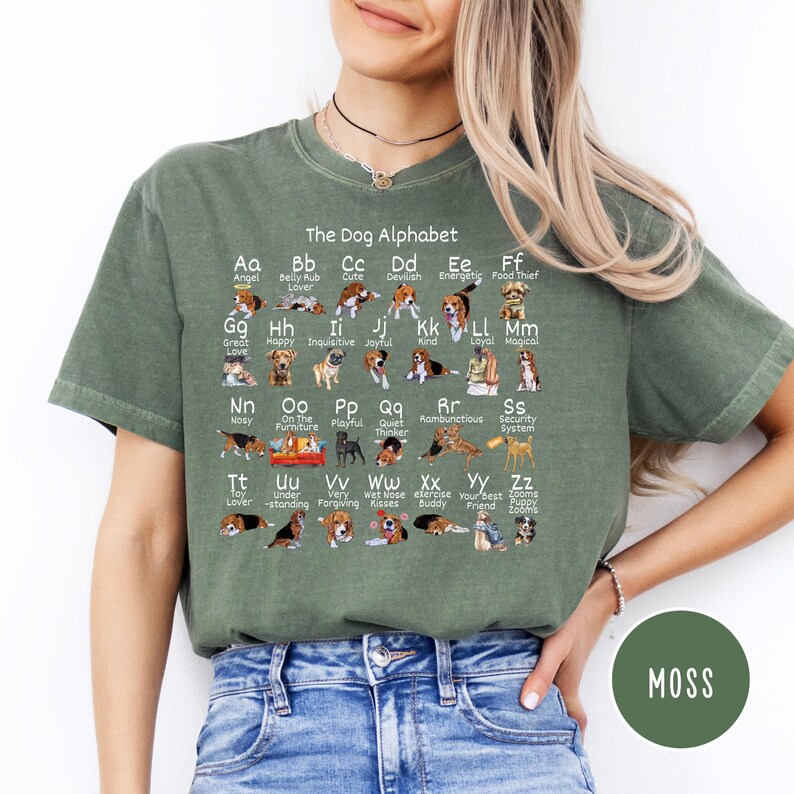May include: A green t-shirt with a graphic of a dog alphabet. The alphabet is printed in black and white with illustrations of dogs for each letter. The text reads "The Dog Alphabet".