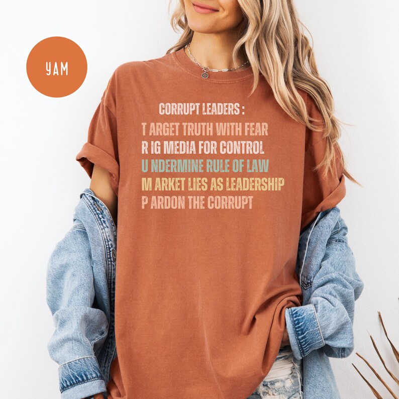 May include: A rust-colored t-shirt with the text "CORRUPT LEADERS: TARGET TRUTH WITH FEAR, RIG MEDIA FOR CONTROL, UNDERMINE RULE OF LAW, MARKET LIES AS LEADERSHIP, PARDON THE CORRUPT." A round orange sticker with "YAM" is in the upper left.