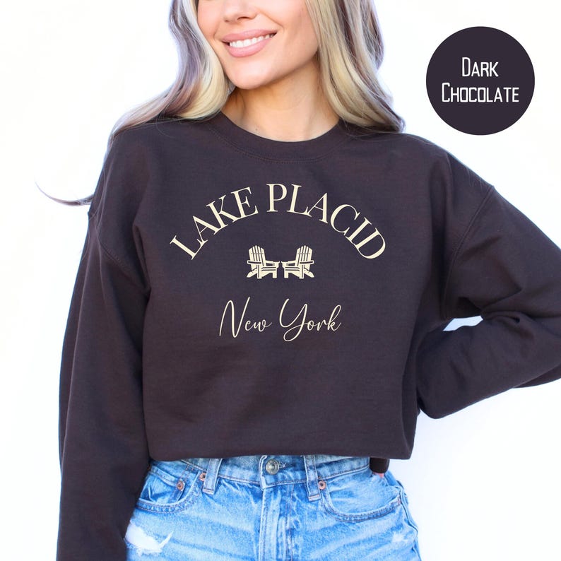 May include: Dark chocolate-colored sweatshirt with the text "Lake Placid New York" in a cream-colored script. The design includes two small chair illustrations. The sweatshirt is a solid color and has a crew neck.