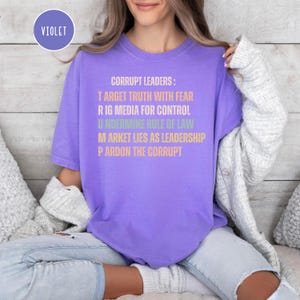 May include: A violet-colored t-shirt with the text "CORRUPT LEADERS: TARGET TRUTH WITH FEAR, RIG MEDIA FOR CONTROL, UNDERMINE RULE OF LAW, MARKET LIES AS LEADERSHIP, PARDON THE CORRUPT." A violet circle with the word "VIOLET" is in the upper left corner.