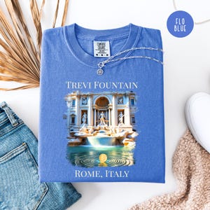May include: A blue t-shirt featuring the Trevi Fountain graphic, with the text "TREVI FOUNTAIN" and "ROME, ITALY". A silver necklace with a pendant rests on the shirt. The shirt is displayed on a white surface, alongside jeans and a scarf.
