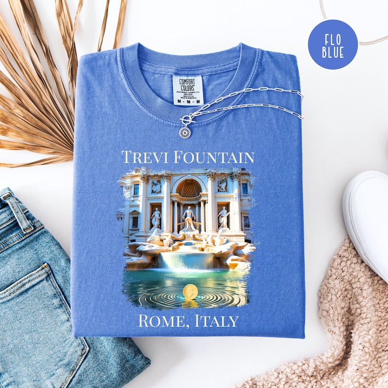 May include: A blue t-shirt featuring the Trevi Fountain graphic, with the text "TREVI FOUNTAIN" and "ROME, ITALY". A silver necklace with a pendant rests on the shirt. The shirt is displayed on a white surface, alongside jeans and a scarf.
