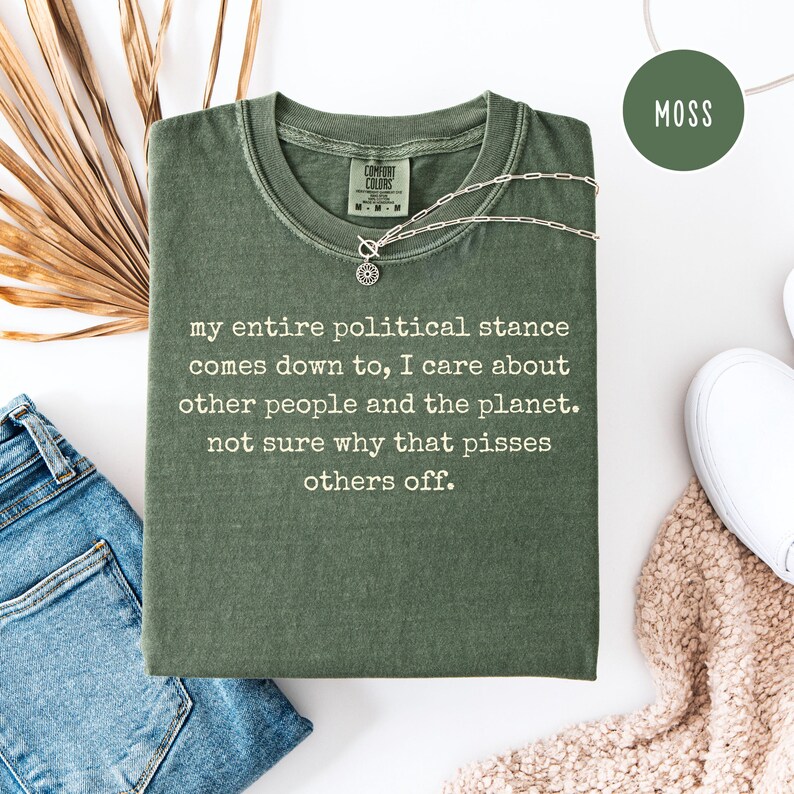 May include: A moss green t-shirt with the text "my entire political stance comes down to, I care about other people and the planet. not sure why that pisses others off." A silver necklace is draped across the shirt.