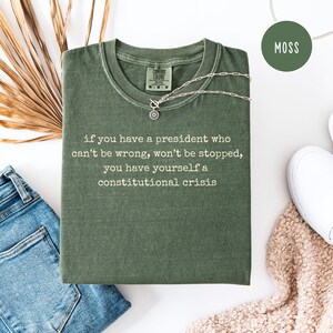 May include: A moss green t-shirt with the text "if you have a president who can't be wrong, won't be stopped, you have yourself a constitutional crisis." A silver necklace with a circular pendant rests on the shirt. The shirt is folded on a white surface.