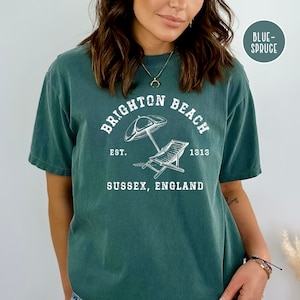 K&ouml;nnte beinhalten: Ein blaugr&uuml;nes T-Shirt mit einem wei&szlig;en Grafikdesign, auf dem "Brighton Beach" mit einem Sonnenschirm und einem Strandstuhl steht. Der Text lautet au&szlig;erdem "Est. 1313" und "Sussex, England".