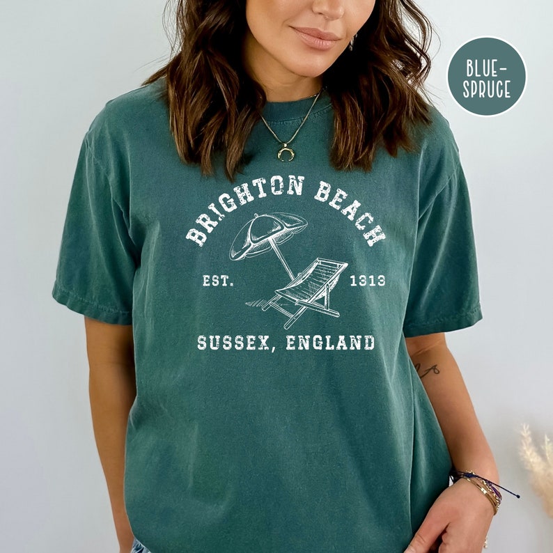 K&ouml;nnte beinhalten: Ein blaugr&uuml;nes T-Shirt mit einem wei&szlig;en Grafikdesign, auf dem "Brighton Beach" mit einem Sonnenschirm und einem Strandstuhl steht. Der Text lautet au&szlig;erdem "Est. 1313" und "Sussex, England".