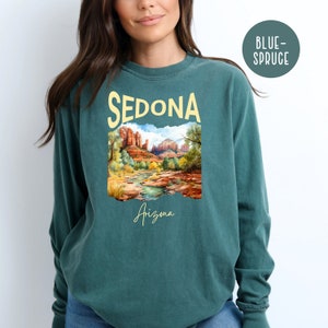 May include: A teal blue long-sleeved t-shirt with a graphic print of a landscape scene with red rock formations and a river. The text "SEDONA" is printed in yellow above the image and "Arizona" is printed in yellow below the image.