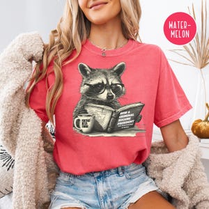 May include: A red t-shirt featuring a raccoon wearing glasses and reading a book titled "How a Criminal Became President." A coffee mug with the text "Foxtrot Delta Tango" is also visible. The shirt has a relaxed fit.
