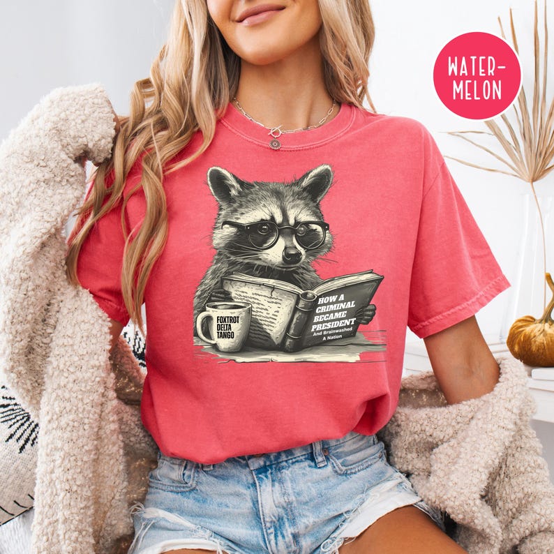 May include: A red t-shirt featuring a raccoon wearing glasses and reading a book titled "How a Criminal Became President." A coffee mug with the text "Foxtrot Delta Tango" is also visible. The shirt has a relaxed fit.