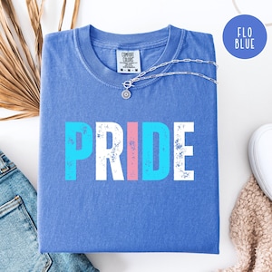 May include: A blue t-shirt with the word "PRIDE" in a distressed font. The letters are in white, blue, and pink. A silver necklace with a flower pendant rests on the shirt. The shirt is folded on a white surface.