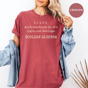 May include: Crimson t-shirt featuring the text "1+1=1 don't overthink it. it's logic, not feelings. BOOLEAN ALGEBRA" in white. The shirt has short sleeves and a solid colour. A denim jacket is worn over the shoulders.
