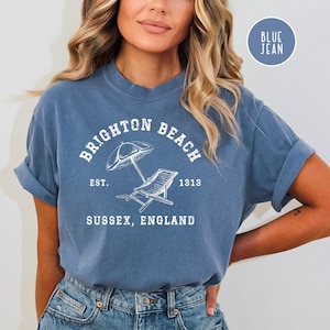 K&ouml;nnte beinhalten: Ein blaues Jeans-T-Shirt mit einem wei&szlig;en Grafikdesign, auf dem "Brighton Beach Est. 1313 Sussex, England" steht, sowie eine Zeichnung eines Strandstuhls und eines Sonnenschirms.