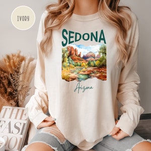 May include: A long-sleeved, ivory-colored sweatshirt with a green graphic print of a landscape scene with red rock formations and a river. The text "SEDONA" is printed in green above the image, and the text "Arizona" is printed in green below the image.