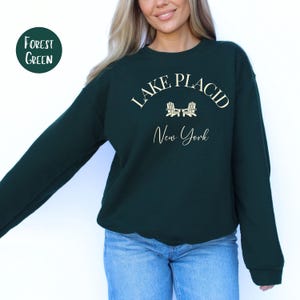 May include: Forest green sweatshirt with the text "Lake Placid New York" in white script. The sweatshirt has a crew neck and long sleeves. A circular label in the upper left corner reads "Forest Green".
