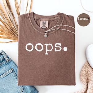 Puede incluir: Una camiseta marrón con la palabra "oops" impresa en blanco en la parte delantera. La camiseta está hecha de una tela suave y cómoda y tiene un corte holgado.