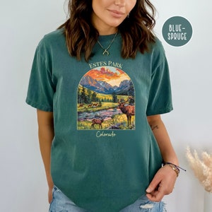 May include: A teal blue t-shirt featuring a graphic print of a mountain landscape with a sunset, a river, and a deer. The text "Estes Park Colorado" is printed on the shirt.