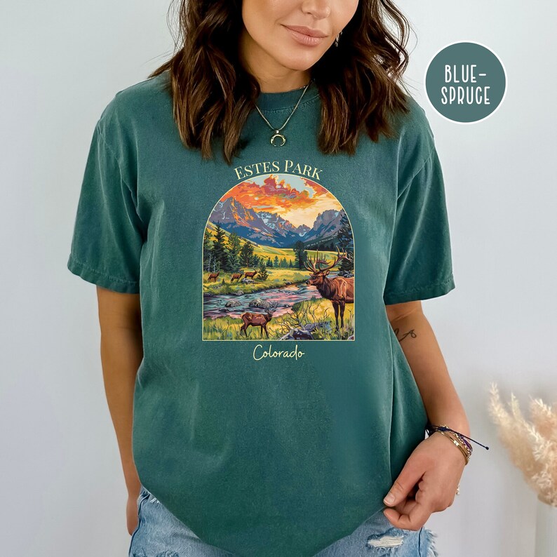 May include: A teal blue t-shirt featuring a graphic print of a mountain landscape with a sunset, a river, and a deer. The text "Estes Park Colorado" is printed on the shirt.
