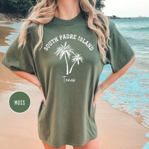 May include: A moss green t-shirt with a white graphic of palm trees and the text "South Padre Island Texas".