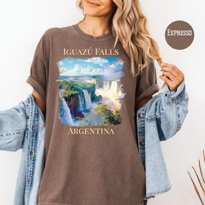Pu&ograve; includere: T-shirt marrone con un'immagine vibrante delle cascate dell'Iguaz&uacute; in Argentina, con un arcobaleno e cielo blu. Il testo "IGUAZ&Uacute; FALLS" e "ARGENTINA" &egrave; stampato sopra e sotto l'immagine. La maglietta ha le maniche arrotolate.