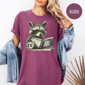May include: Berry-colored t-shirt featuring a raccoon wearing glasses and reading a book titled "How a Criminal Became President." A coffee mug with the text "Foxtrot Delta Tango" sits beside the book. The shirt has rolled-up sleeves.